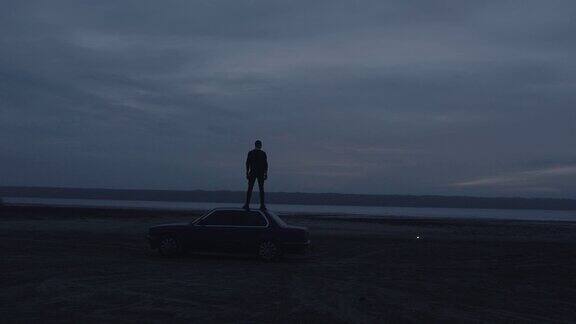 手持电影黑暗的年轻人站在车与红色信号燃烧信号弹在海滩附近的水