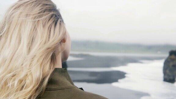 年轻的金发女人站在海边看着巨怪脚趾岩石在冰岛在阴天的景象