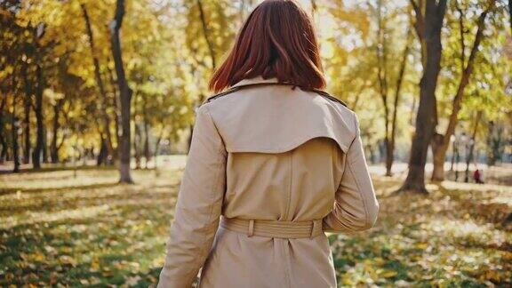 一名年轻女子微笑着走在秋天的城市公园温暖阳光的日子黄澄澄的树木跟踪镜头慢动作后视镜