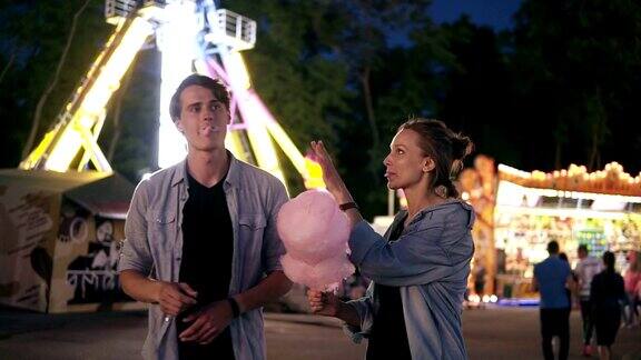 年轻的时髦夫妇晚上一起在游乐园玩互相喂粉红棉花糖前视图