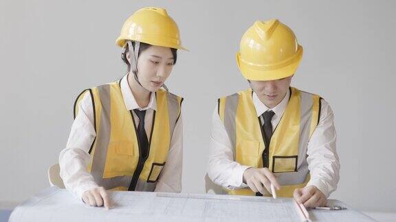 两个亚洲工程师穿着安全背心和安全帽正在检查建筑图纸讨论设计方案