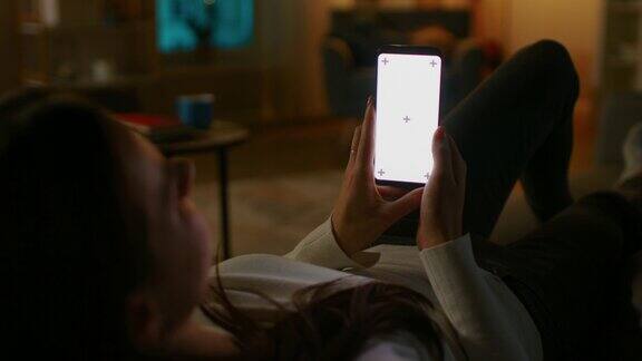 晚上在家:年轻女子坐在沙发上使用白色模拟屏幕智能手机女孩使用ChromaKey手机上网在社交网络上发帖