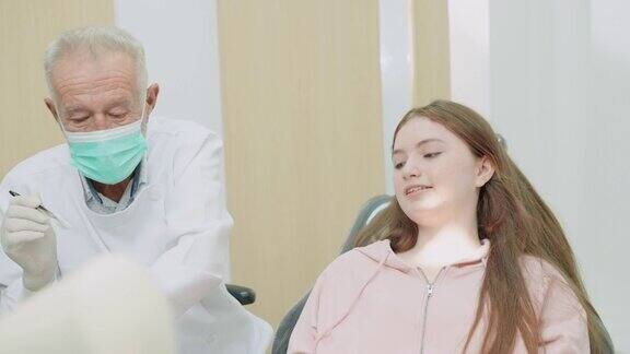 老牙医为十几岁的少女病人补牙后在灯光下检查牙齿并看牙洞少女坐在牙科单元上无所畏惧侧视