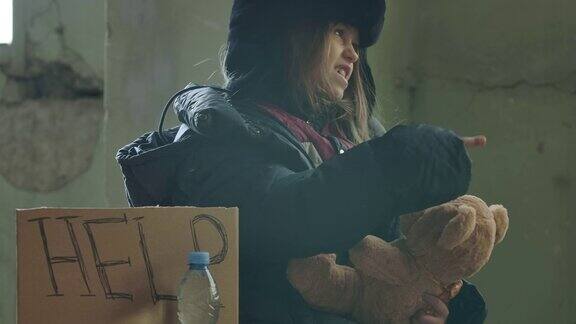 一名叙利亚难民的肖像穿着脏衣服坐在“帮助”纸板上和她的泰迪熊说话