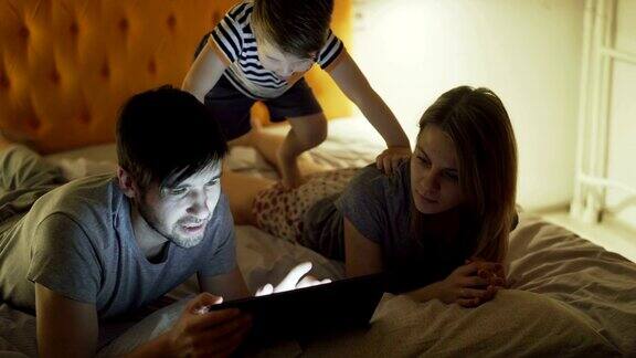一个幸福的家庭小儿子躺在家里的床上在睡觉前用平板电脑浏览社交媒体