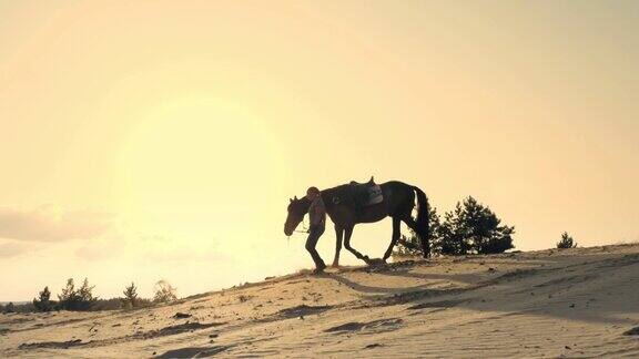 骑马女骑手的剪影牵着一匹马沿着地平线高耸的沙山在夕阳西下时天空的背景和阳光的光线背光