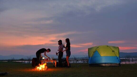 场景一群青少年亚洲人正在一起享受帐篷前的篝火和看日落附近的湖在暑假他们是快乐的有乐趣的假期放松的时刻青少年自然户外日常生活