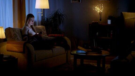 在晚上美丽的年轻女人放松在沙发上在她舒适的客厅她一边用智能手机一边看电视
