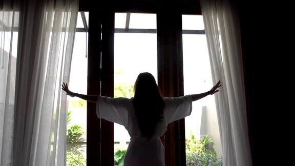 年轻女子穿着浴袍从床上站起来拉开窗帘伸懒腰站在家里靠近窗户的地方早上醒来