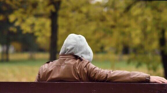一个戴着兜帽的不明身份男子坐在秋天公园的长椅上