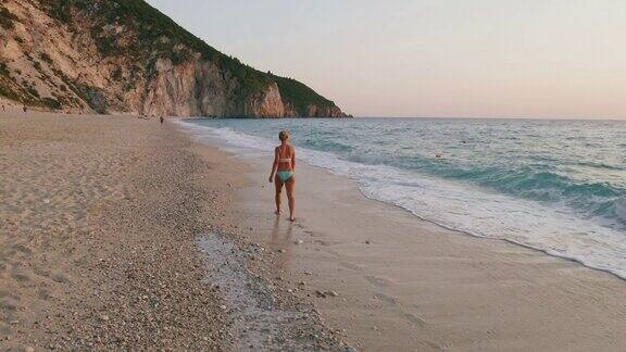 空中低空跟随成年妇女享受美丽的米洛斯海滩的Lefkada爱奥尼亚岛希腊夕阳金色的阳光和原始的海浪打破了蓝绿色的大海在岸边