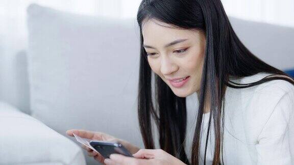 年轻的亚洲女子躺在客厅的沙发上用智能手机上的银行卡通过互联网进行网上银行支付用信用卡在手机上网购