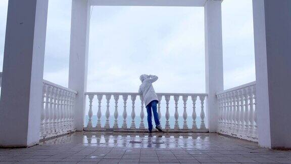在美丽的阳台上穿着白色夹克的女人欣赏着海景后视图