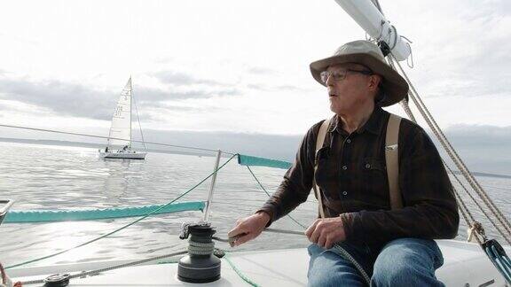 华盛顿普吉特海湾一名六十多岁的男子在他的帆船上用绞车拉紧绳子而他的船员坐在甲板上看风景