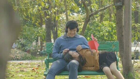 小情侣在公园的长椅上放松 女孩倚在男友的大腿上看书