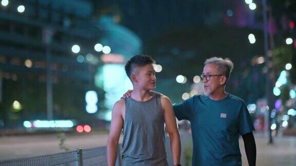 亚洲华人活跃的老人和他的儿子在晚上一起跑步锻炼后散步聊天城市