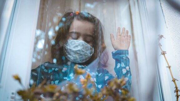 看起来很伤心的小女孩戴着防护面具透过窗户看因为她不被允许出去玩