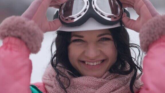 迷人的快乐的女人在镜头与牙齿微笑戴上滑雪镜在慢动作的特写肖像快乐兴奋的高加索滑雪者摆姿势在户外冬季度假村