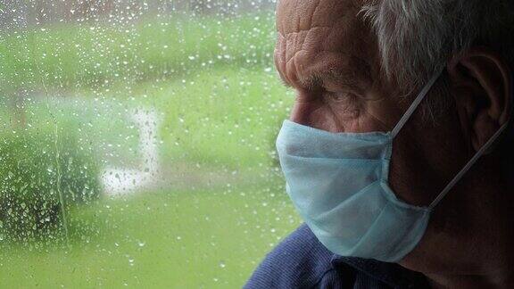 下雨时戴着医用防护口罩的悲伤老人望着窗外的街道
