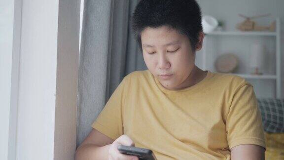 亚洲男孩坐在家里靠窗的地方用智能手机看电影或视频生活理念