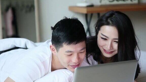 亚洲华人夫妇拥抱、亲吻、微笑早上在床上用笔记本电脑上网