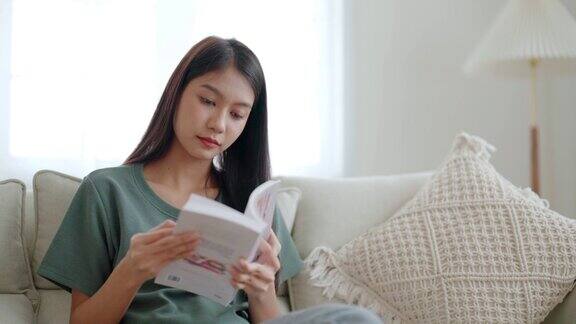 快乐的年轻亚洲女人放松在舒适的沙发在家里读书和坐在沙发上