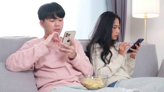 年轻的亚洲夫妇坐在沙发上玩他们的手机他们在专注于在线内容的同时忽略了彼此