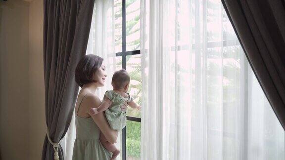 快乐的亚洲妇女的肖像抱着婴儿靠近窗户看向别处的家4个月大的婴儿抱在父母怀里母女关系妈妈抱着婴儿在卧室里