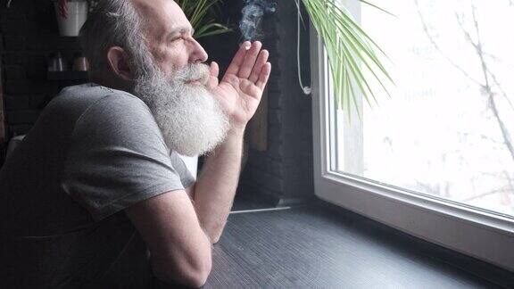 一位留着胡子、头发花白的老人望着窗外然后在厨房里抽烟