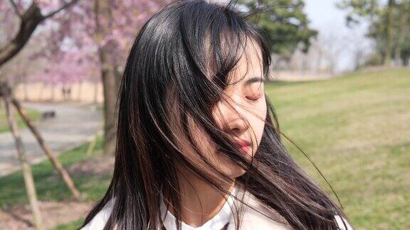 美丽的中国年轻女子在白色外套与粉红色的樱花背景闭上眼睛享受微风吹她的黑色长发美丽的春天4k慢镜头