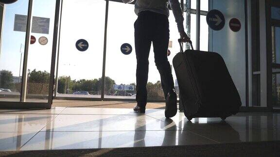 年轻商人带着行李从机场走出来的脚步声一名男子穿过玻璃自动门来到街上用轮子推着行李箱太阳耀斑在背景旅行或旅行概念慢动作特写后视图