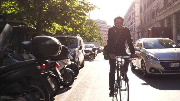 在阳光明媚的日子里年轻的潮男骑着自行车在罗马市中心一条有树的道路上戴着墨镜
