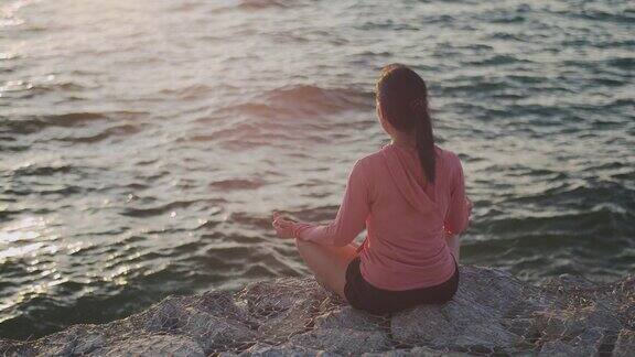亚洲美丽的女孩运动女孩训练冥想坐在石头海边在日落