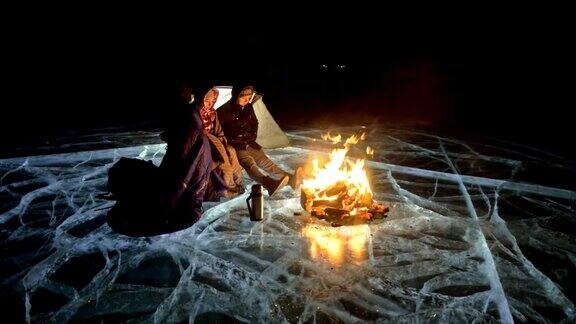 三个旅行者在夜间的冰面上着火营地在冰上帐篷紧挨着火贝加尔湖附近有一辆车人们围着篝火取暖穿着睡袋