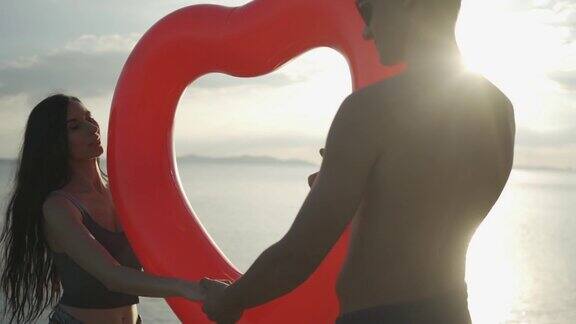 男人和女人的剪影拿着一个心脏游泳环在海滩上走在太阳下的日落