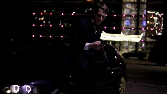 穿着西装的商人在一辆豪华行政轿车旁用着智能手机停车场有个大窗户背景灯光