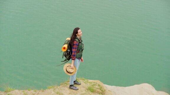 从上面跟踪拍摄的一位年轻的女旅行者背着背包走下山走向大海摘下草帽欣赏美丽的风景在刮风的日子