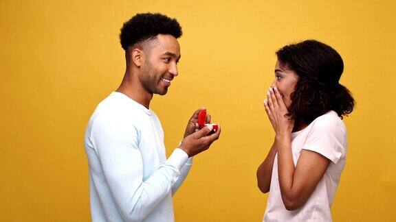 英俊的年轻非洲男子向他的女朋友求婚戒指在一个盒子孤立的黄色背景