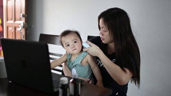 远程医疗亚洲华人母亲用体温计检查她的男婴的体温同时用笔记本电脑在线咨询她的儿科医生