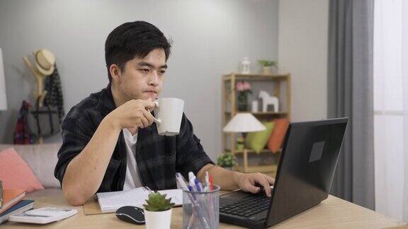 一名亚洲男性自由职业工人一边喝茶一边在家里的电脑上继续工作一边点头