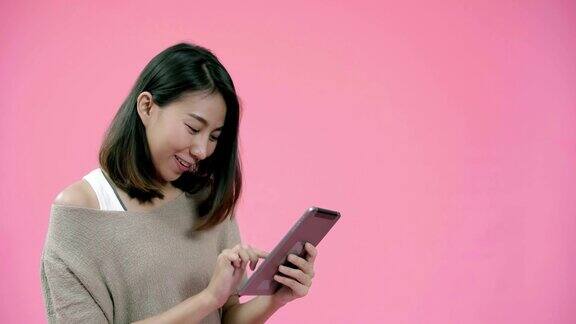 年轻的亚洲女性用平板电脑查看社交媒体在粉色背景的工作室拍摄下穿着休闲服装开心地微笑快乐的微笑可爱的快乐的女人庆祝成功