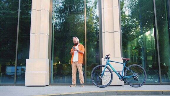在一个现代化的办公中心附近一个满脸胡须、手里拿着电话的快乐男人一个骑自行车的年轻人拿着智能手机站在楼前