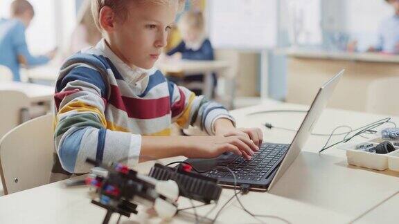 一名男生将电缆连接到一个小机器人上用笔记本电脑为机器人工程课编写软件小学科学教室有天赋的聪明的孩子与技术工作