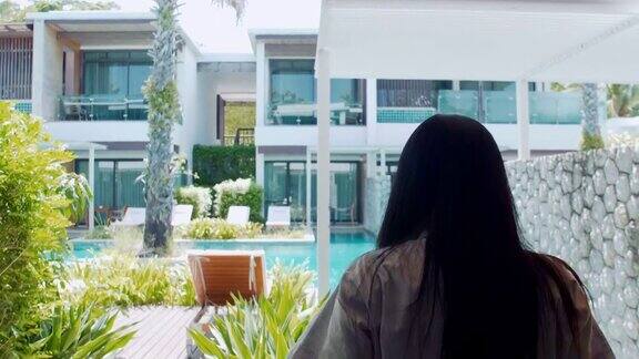 一位亚洲年轻女子拉开窗帘穿着浴袍的女士打开酒店房间的白色窗帘透过窗户欣赏幸福女人们拉开窗帘走向酒店房间的游泳池