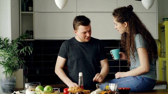一对快乐的年轻夫妇在家里的厨房里做饭和聊天帅哥一边喂女友一边切菜做早餐