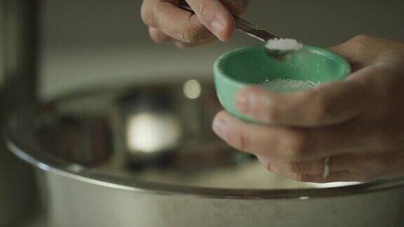 一个年轻女人的手用一个小金属量勺从搅拌碗旁边的小碗里舀盐