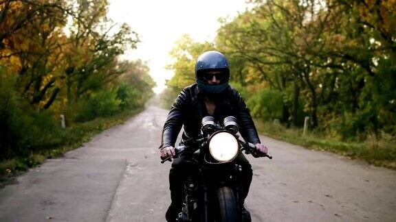正面的观点一个年轻人戴着头盔戴着太阳镜和皮夹克骑摩托车在柏油路在秋天路的周围长着棕色和黄色叶子的树