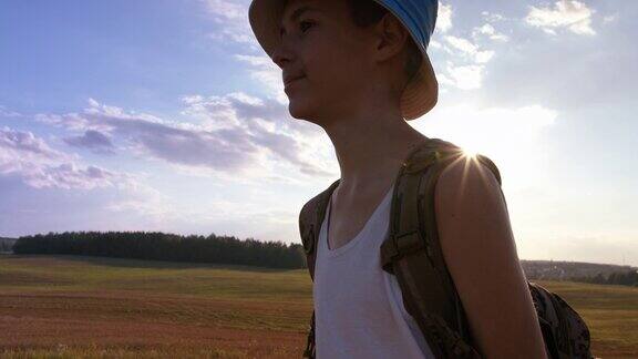 夏天日落时分背着背包的少年旅行者在一座小山的山顶上走过田野