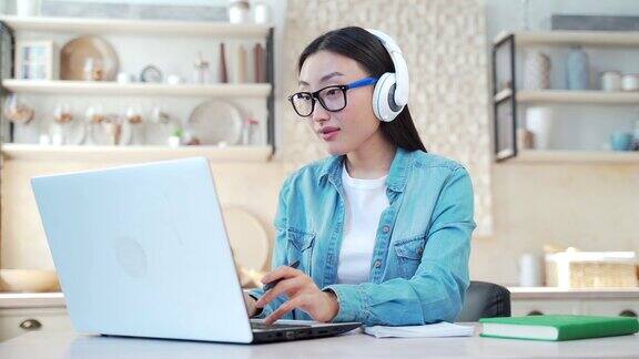 年轻的亚洲女子戴着耳机在笔记本电脑上打字坐在客厅的桌子上写笔记