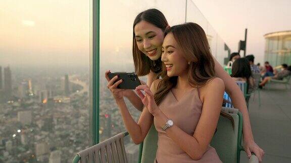 日落时分4K的亚洲女性朋友在摩天大楼的屋顶餐厅一起用智能手机自拍